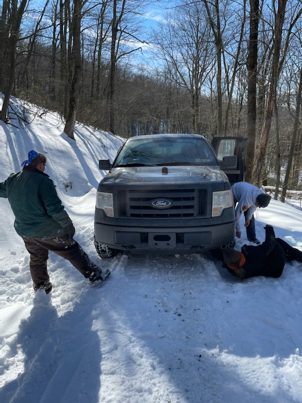 truck of snow covered road with people putting chains on tires while one stands to watch